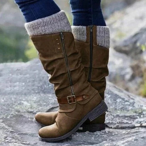 Eine Nahaufnahme eines braunen orthopädischen Stiefels mit lässigem Design und Strickbund, der von einer Person getragen wird, die auf felsigem Boden steht und sein stilvolles Aussehen und seine Komfortmerkmale hervorhebt.